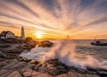 Maine rocky coastline at golden sunset, lighthouse on cliffs, waves crashing, lobster boat offshore