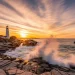 Maine rocky coastline at golden sunset, lighthouse on cliffs, waves crashing, lobster boat offshore